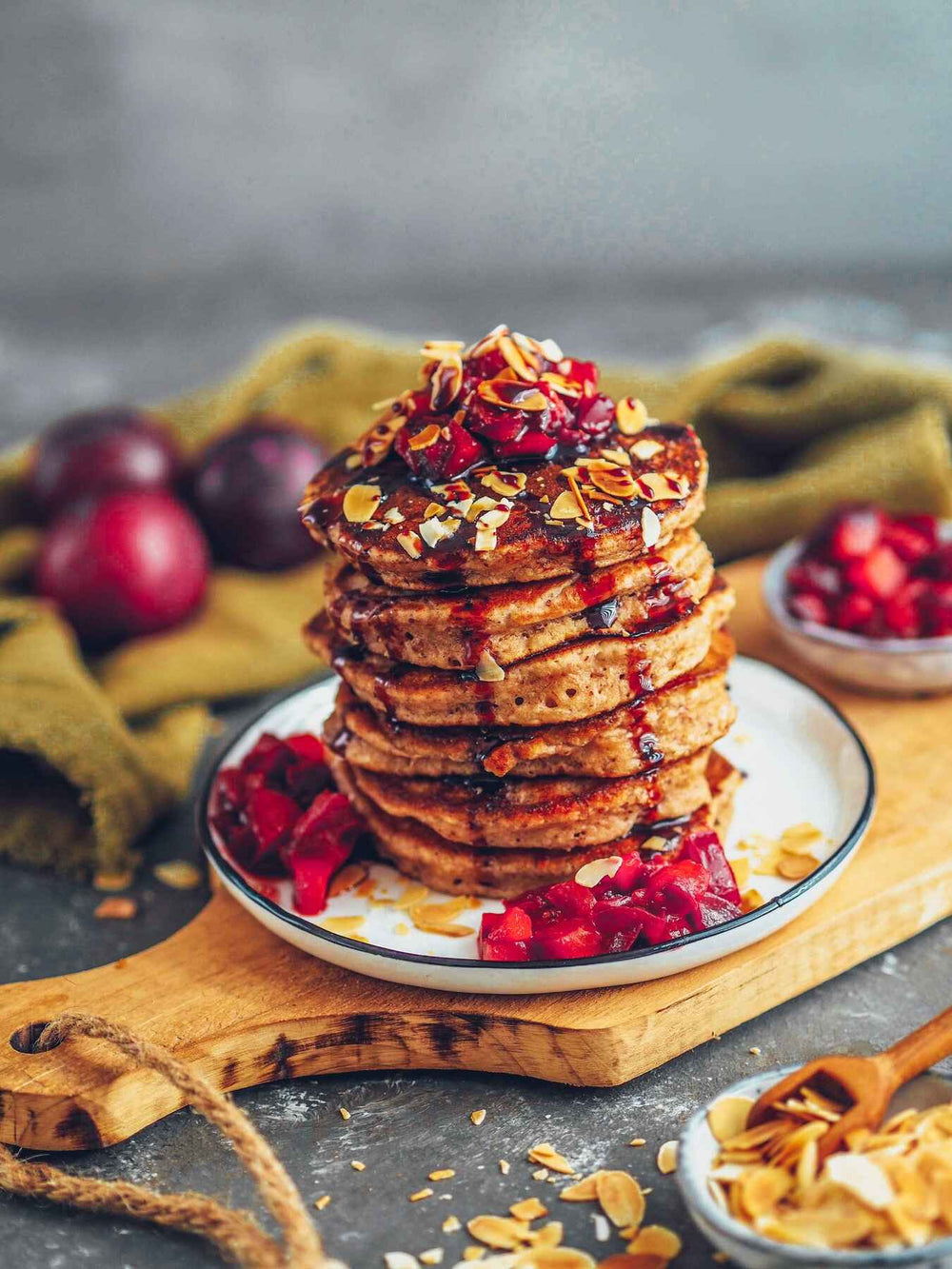 Ein hoher Stapel Pfannkuchen mit gehackten Nüssen und Obstkompott liegt auf einem weißen Teller. Weitere Früchte und Nüsse liegen auf dem Schneidebrett verstreut, mit Pflaumen und einem grünen Tuch im unscharfen Hintergrund.