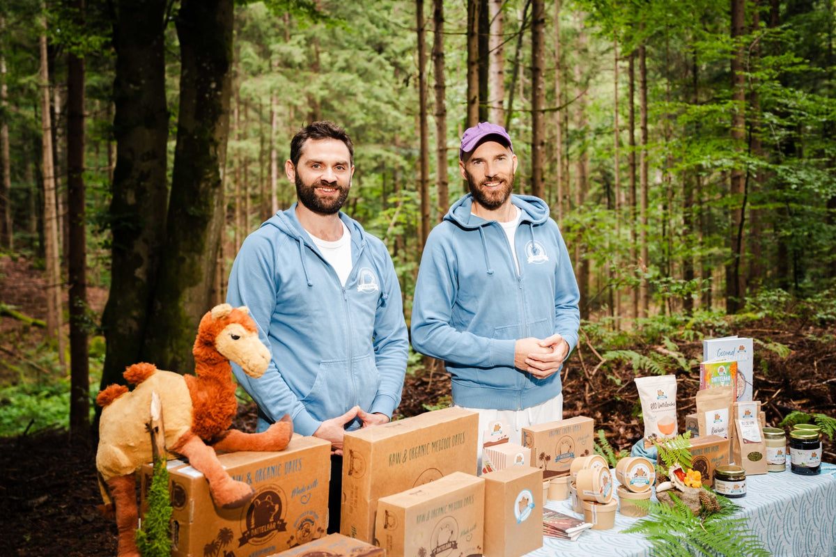 Zwei Männer in blauen Kapuzenpullovern stehen hinter einem Tisch mit Lebensmitteln und einem Plüschkamel in einer Waldlandschaft. Der Tisch ist mit Schachteln, Gläsern und verpackten Waren bedeckt. Hohe grüne Bäume umgeben die Szene.