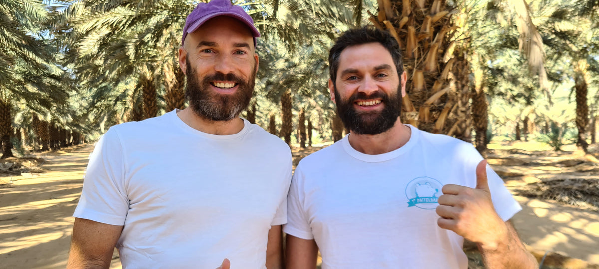 Zwei lächelnde Männer mit Bärten stehen dicht beieinander im Freien unter hohen Palmen. Beide tragen weiße T-Shirts, einer hat eine kastanienbraune Mütze auf und der andere zeigt den Daumen nach oben. Helles Sonnenlicht fällt durch die Bäume.