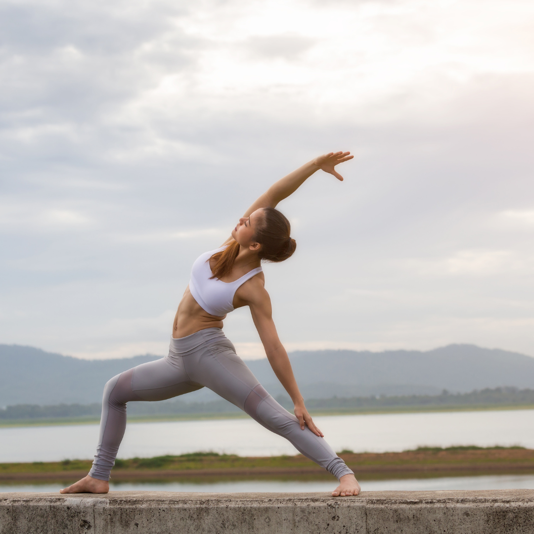 Eine Frau in Sportkleidung nimmt im Freien auf einer Betonfläche eine Yogastellung ein und streckt dabei einen Arm über den Kopf. Im Hintergrund sind ein ruhiger See und Berge unter einem bewölkten Himmel zu sehen.
