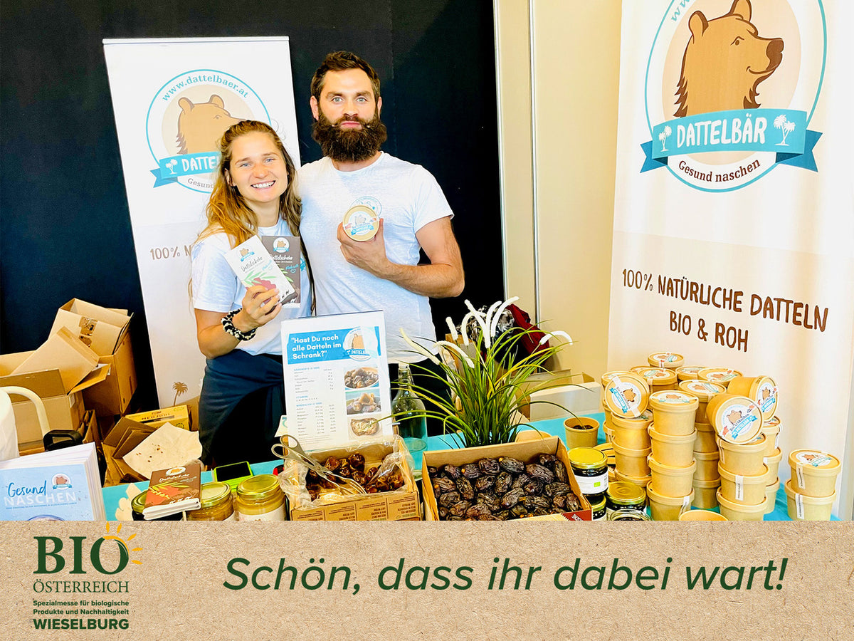 Eine lächelnde Frau und ein bärtiger Mann stehen an einem Stand, der Dattelsnacks verkauft. Der Stand zeigt ein Bärenlogo, Produktdisplays und ein Banner mit der Aufschrift "DATTELBÄR". Der Text darunter lautet: "Schön, dass ihr dabei wart!".