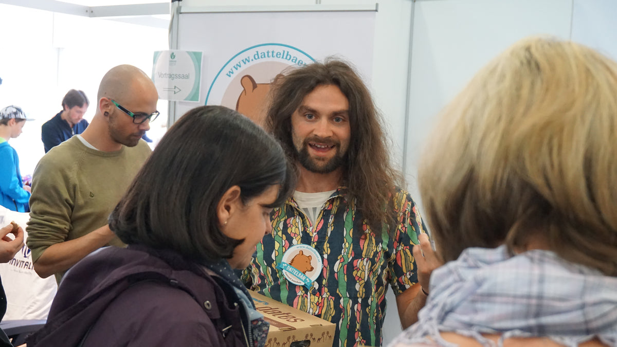 Ein lächelnder Mann mit langen Haaren, der ein bunt gemustertes Hemd trägt, steht an einem Stand mit einer Schachtel in der Hand und unterhält sich mit Leuten bei einer Veranstaltung in einer Halle. Andere Teilnehmer sind im Hintergrund zu sehen.
