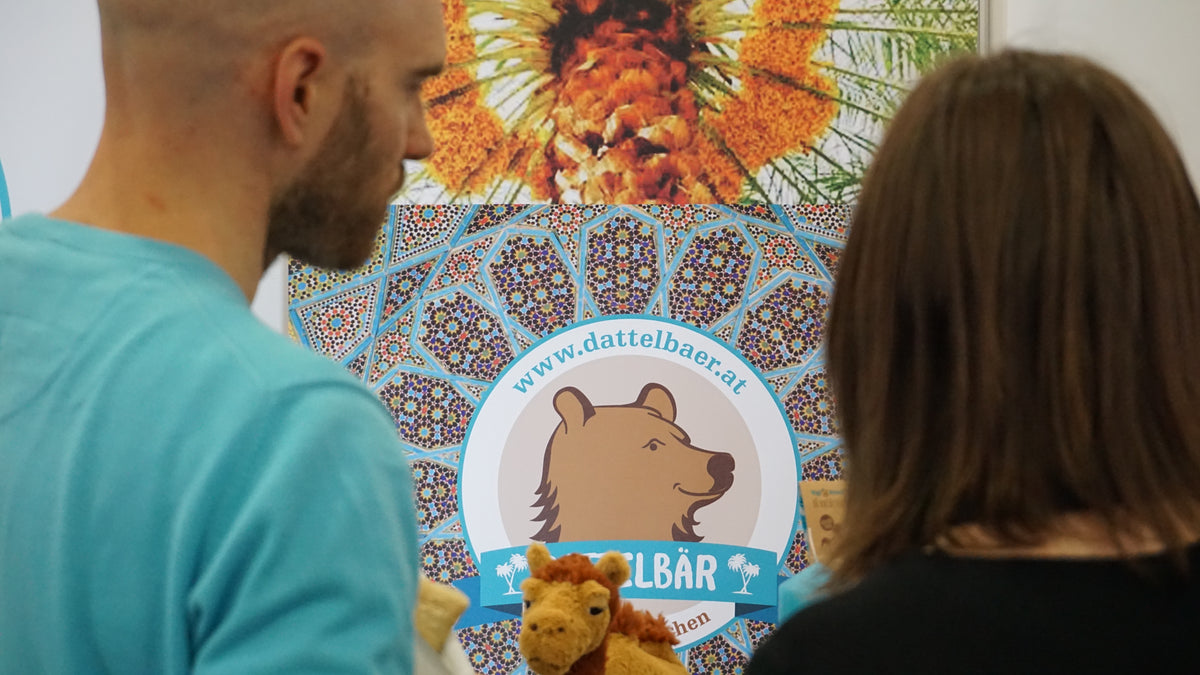 Zwei Personen stehen vor einem farbenfrohen Display mit einem Logo mit einem Bären, verschlungenen Mustern und der Website www.dattelbaer.at. Im Vordergrund ist auch ein Kamel aus Plüsch zu sehen.