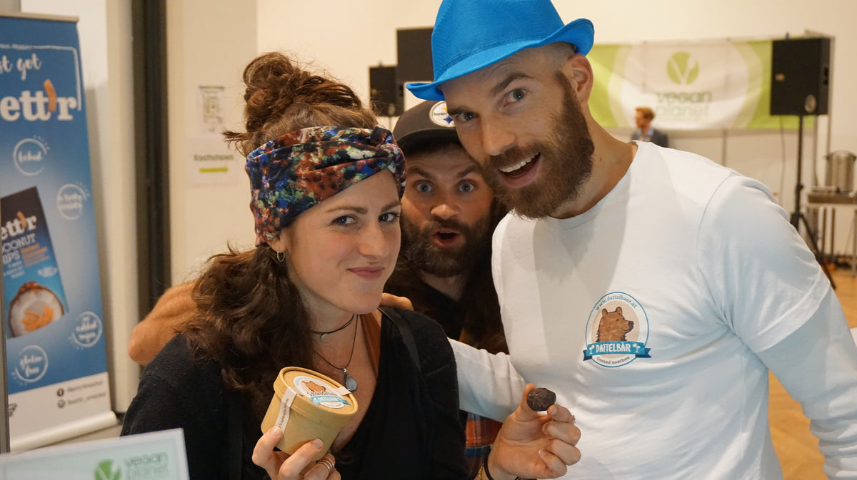 Drei lächelnde Menschen posieren bei einer veganen Veranstaltung in einem Innenraum. Die Frau vorne hält einen Becher mit einem veganen Produkt, während die beiden Männer hinter ihr, von denen einer einen blauen Hut trägt, sich spielerisch anlehnen. Im Hintergrund sind Schilder und Transparente zu sehen.