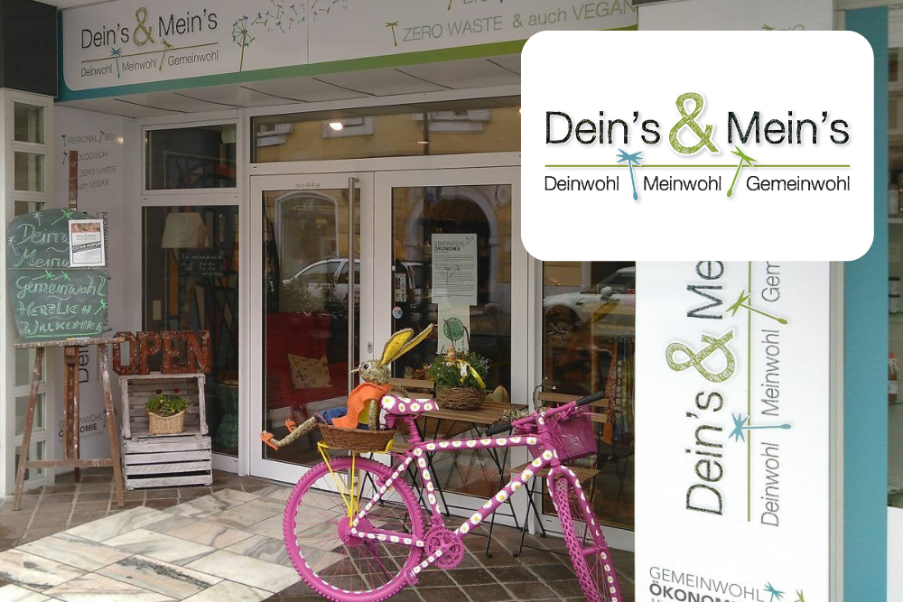 Ein veganer, abfallfreier Laden namens "Dein's & Mein's" hat ein rosa Fahrrad mit Körben voller Produkte vor seinem Eingang stehen. Eine Kreidetafel und Topfpflanzen stehen neben den Glastüren. Das Ladenschild und das Logo sind sichtbar.