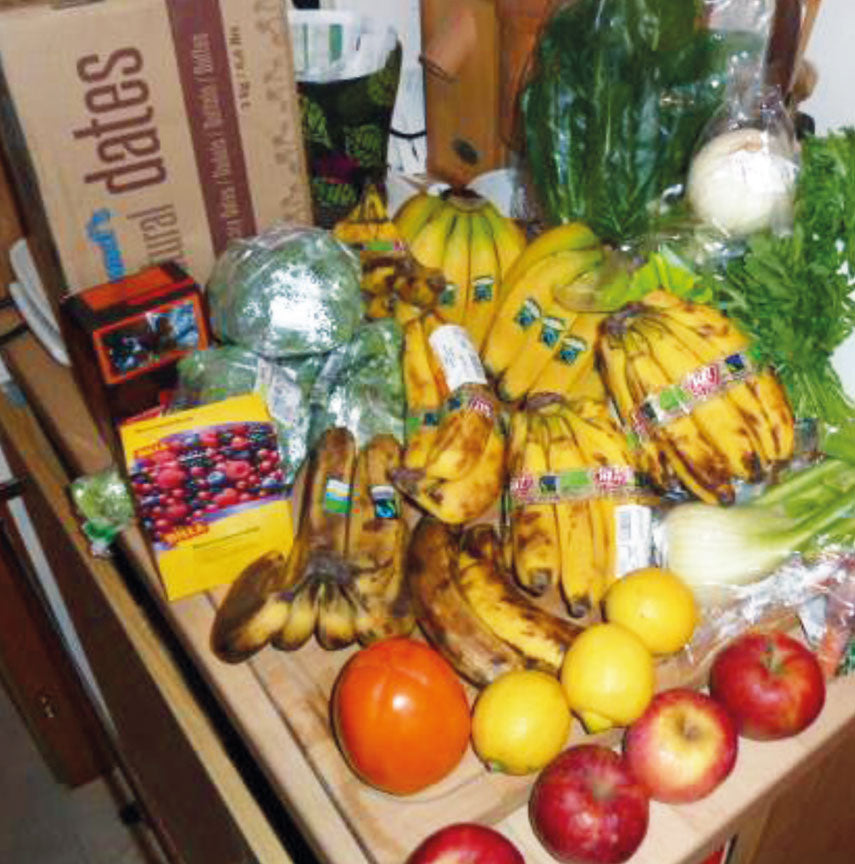 Eine Küchentheke, gefüllt mit verschiedenen Lebensmitteln, darunter Bananen, Äpfel, Zitronen, Salat, Kohl, Tomaten, Sellerie, abgepackte Datteln und andere Obst- und Gemüsesorten sowie einige verpackte Lebensmittel.