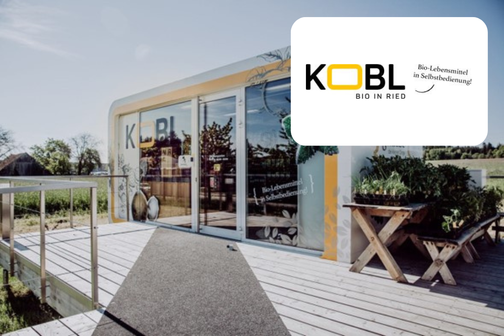 Ein modernes, kleines Gebäude mit großen Glastüren und einer Holzterrasse ist von Grünflächen unter einem klaren Himmel umgeben. Das Logo von KOBL BIO IN RIED wirbt für biologische Selbstbedienungsprodukte.