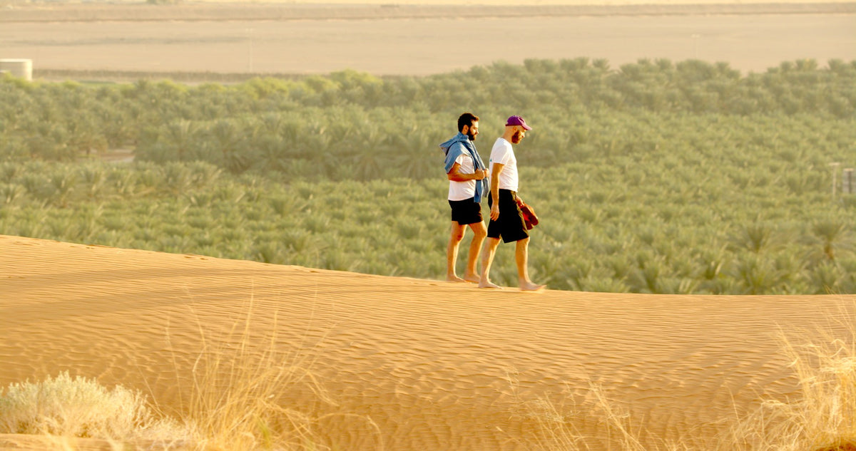 Zwei Menschen laufen barfuß über goldene Wüstensanddünen und tragen dabei ihre Schuhe. Im Hintergrund ist ein üppiger grüner Palmenhain zu sehen, der sich von dem trockenen Sand abhebt. Beide tragen kurze Hosen und T-Shirts.