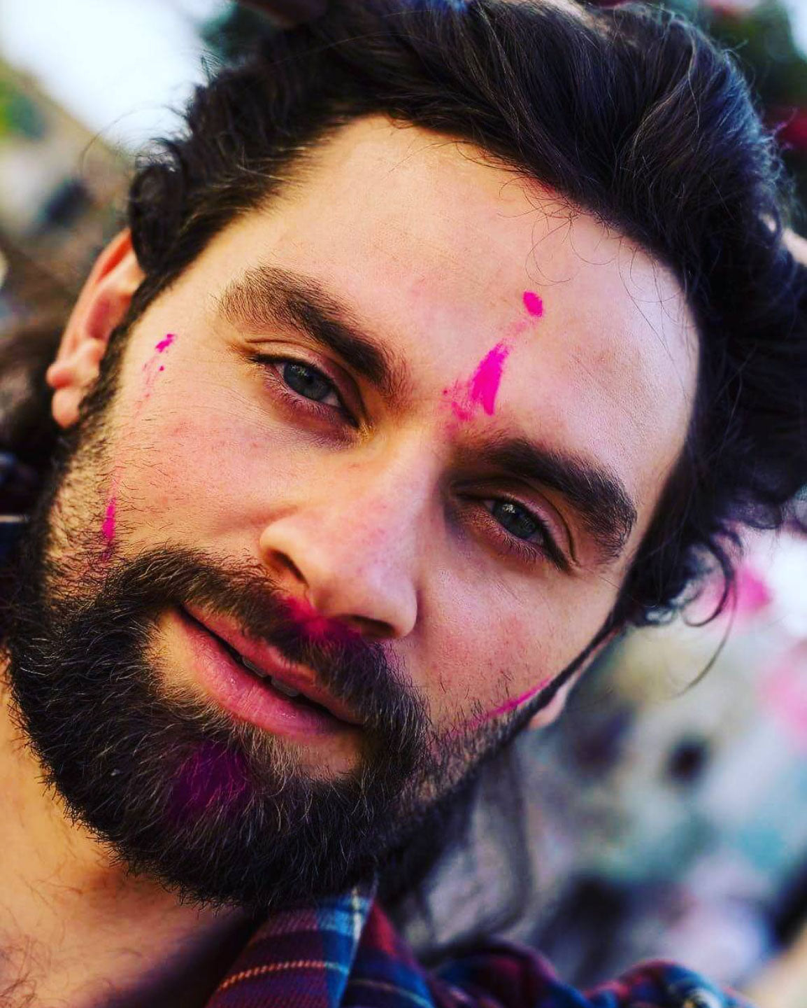 Ein Mann mit einem Bart und langen Haaren lächelt leicht. Er hat Schlieren von rosa Farbpulver im Gesicht, was darauf hindeutet, dass er ein Fest wie Holi feiert. Der Hintergrund ist unscharf.