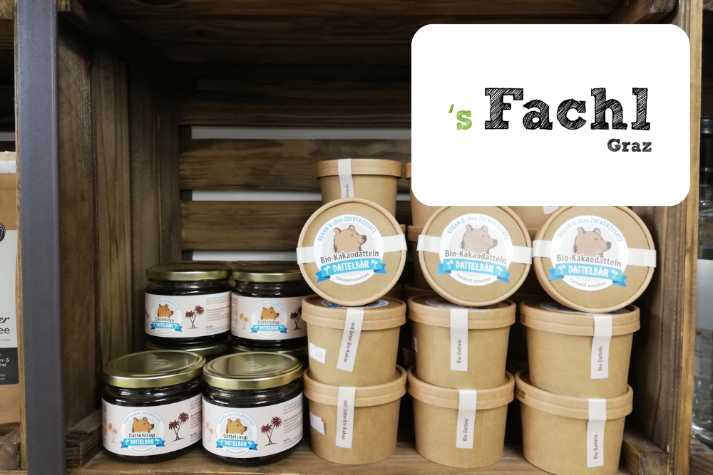 Auf einem Holzregal stehen Tiegel und braune Pappbecher mit Bio-Kakaocreme. Ein weißes Schild mit schwarzer und grüner Schrift zeigt in der oberen rechten Ecke s Fachl Graz.