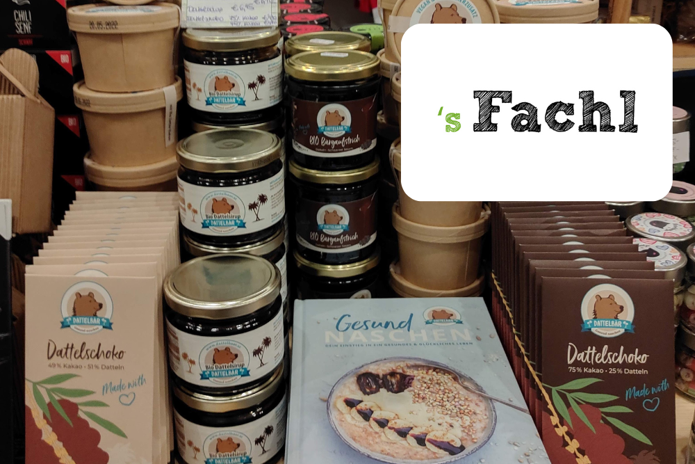 Auslage von Schokoriegeln, Aufstrichgläsern und verpackten Snacks in einem Ladenregal. Zu sehen sind ein Buch mit dem Titel Gesund Naschen und ein weißes Schild mit s Fachl in grüner und schwarzer Schrift.