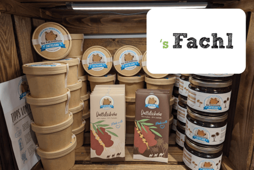 In einem Holzregal stehen Naturprodukte in braunen Papier- und Kartonverpackungen, darunter Gläser, Becher und Snackriegel mit der Aufschrift Dattelschoko. Auf einem Schild darüber steht in fetter schwarzer und grüner Schrift "s Fachl".