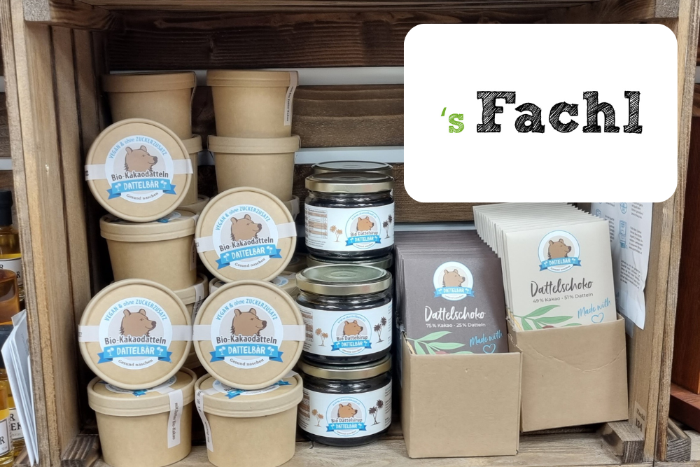 In einer Holzkiste stehen Gläser und Pappbecher mit Etiketten, die einen Bären und den Text Bio Kakao Creme UNTEILBAR zeigen. Daneben stehen Päckchen mit der Aufschrift Dattelschnitte. Auf einem weißen Schild steht s Fachl.