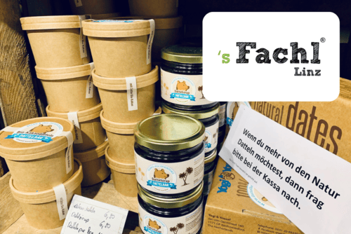 Eine Auslage mit Naturkostprodukten im s Fachl Linz, darunter braune Papierwannen, Gläser mit Tieretiketten und eine Schachtel mit der Aufschrift Naturdatteln. Ein Schild in deutscher Sprache fordert die Kunden auf, sich an der Theke nach weiteren natürlichen Datteln zu erkundigen.