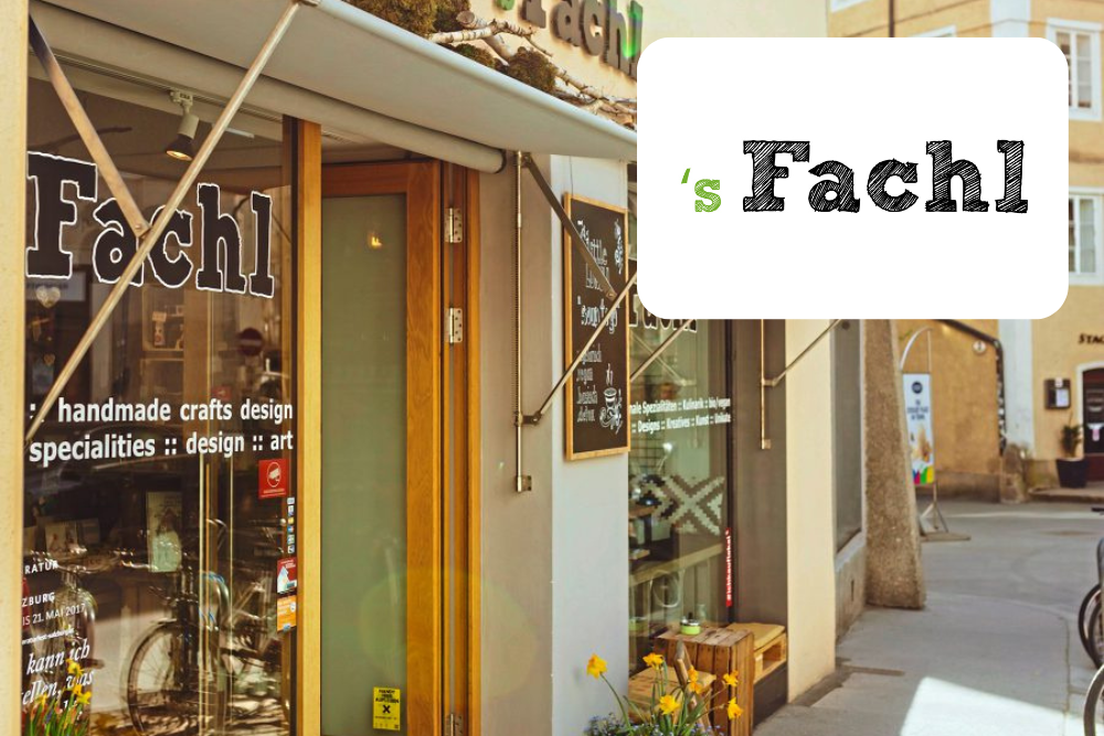 Eine Schaufensterfront mit großen Fenstern, auf denen der Name "Fachl" steht. Auf der Tür und den Fenstern ist ein Text zu sehen, der für handgefertigtes Kunsthandwerk und Design wirbt. Im Hintergrund ist eine Straßenszene mit Gebäuden und Menschen zu sehen.