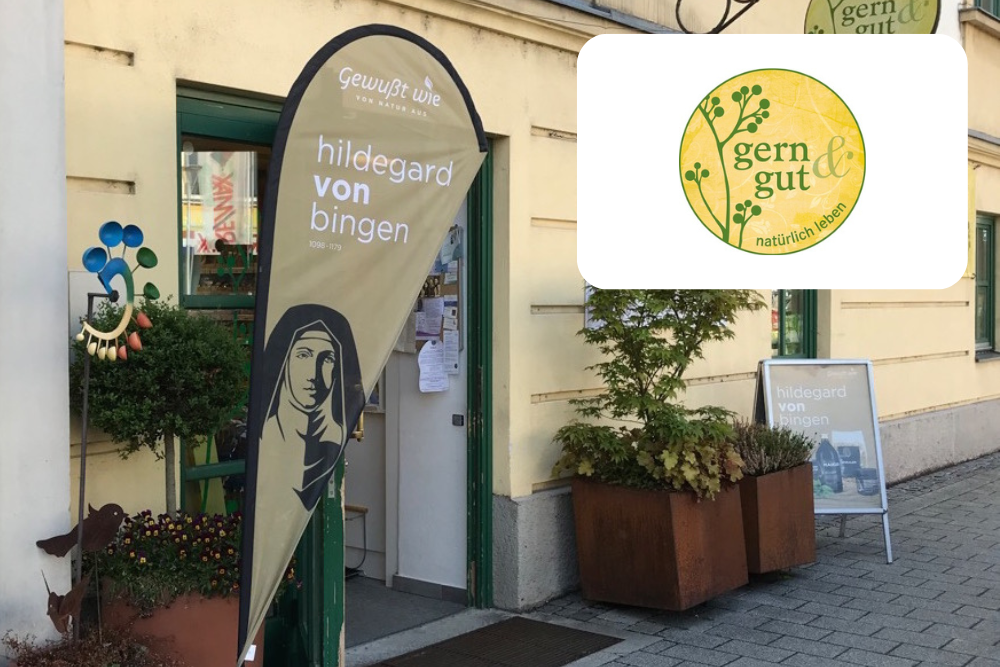 Eine Schaufensterfront mit einer Fahne, auf der Hildegard von Bingen und ihr Porträt abgebildet sind, neben einer grünen Tür mit Pflanzen draußen. Das Logo gern gut mit gelber und grüner Schrift und Beeren ist in der oberen rechten Ecke abgebildet.