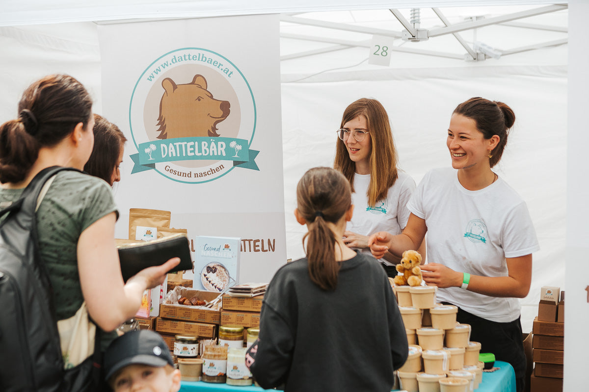 Zwei Frauen stehen hinter einem Marktstand für Dattelbär, lächeln und sprechen mit Kunden. Der Stand zeigt Gläser, Produkte und ein Bärenlogo auf einem Transparent. Mehrere Personen, darunter auch Kinder, stehen vor dem Stand.