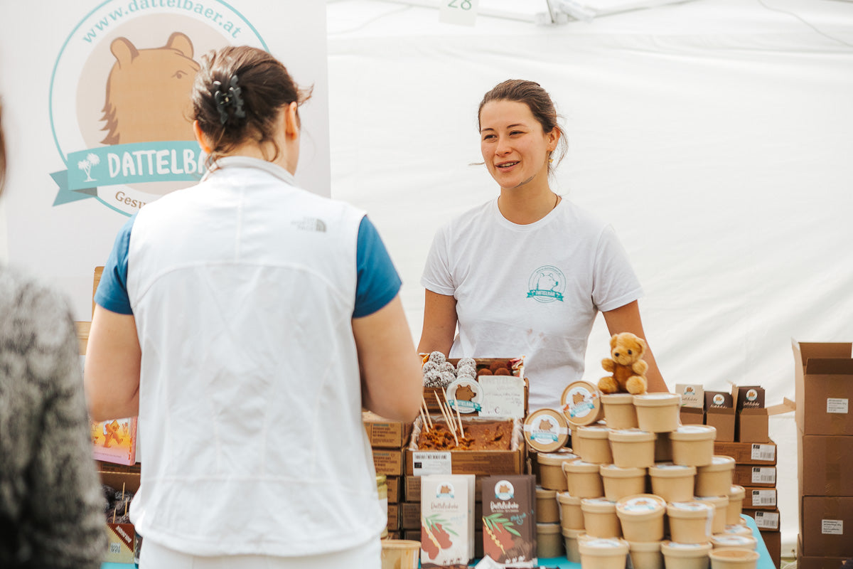 Eine Frau hinter einem Marktstand lächelt und spricht mit einem Kunden. Der Stand bietet Gläser, Snackbars und einen kleinen Teddybären an. Das Standschild zeigt einen Bären und die Aufschrift DATTELBÄR.