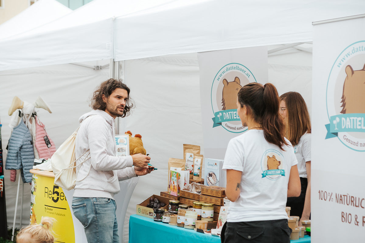 Ein Mann spricht mit zwei Frauen an einem Marktstand, der Snacks verkauft. Die Frauen tragen weiße Hemden mit einem Logo, das einen Bären zeigt. Auf dem Tisch liegen verschiedene verpackte Produkte unter einem weißen Zeltdach.