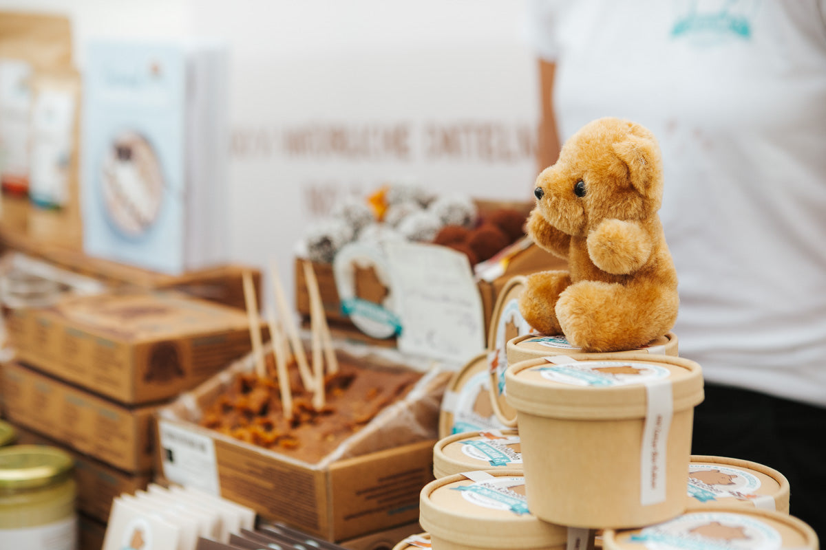 Ein kleiner brauner Teddybär sitzt auf gestapelten Eimern mit Lebensmitteln an einem Marktstand. Im Hintergrund sind Backwaren, verpackte Leckereien und eine Person in einem weißen Hemd zu sehen.