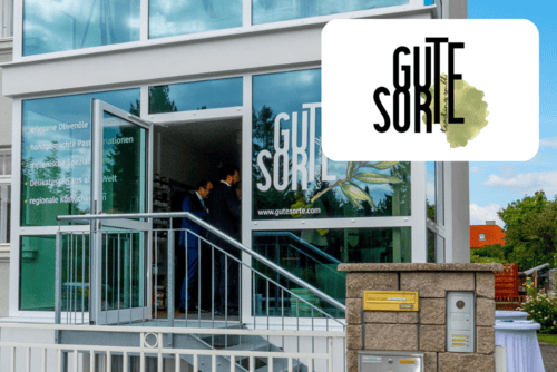 Ein modernes Schaufenster mit großen Glasfenstern und der Aufschrift GUTE SORTE. Zwei Personen stehen direkt vor dem Eingang. In der Ecke ist das GUTE SORTE Logo auf weißem Hintergrund zu sehen.