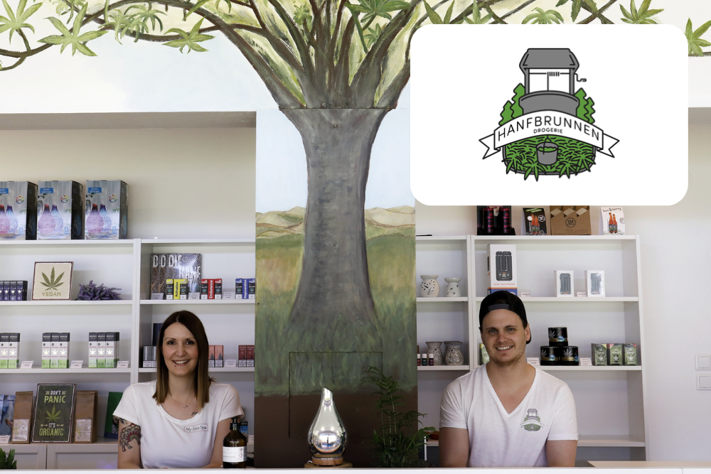 Zwei lächelnde Menschen stehen hinter dem Tresen eines Ladens mit Hanfprodukten in den Regalen. An der Wand hinter ihnen ist ein Wandbild mit einem Baum gemalt. In der Ecke befindet sich ein Logo mit der Aufschrift HANFBRUNNEN mit Blättern und einem Brunnen.