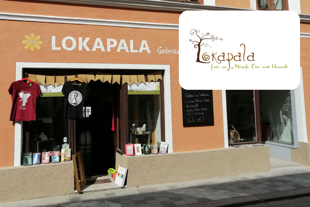 Ein kleiner Laden namens LOKAPALA mit orangefarbenen Wänden und einer Auslage mit Büchern, T-Shirts und Kleinigkeiten im Außenbereich. Am Eingang hängen eine Kreidetafel und ein Banner; das Logo des Ladens ist in der oberen rechten Ecke zu sehen.