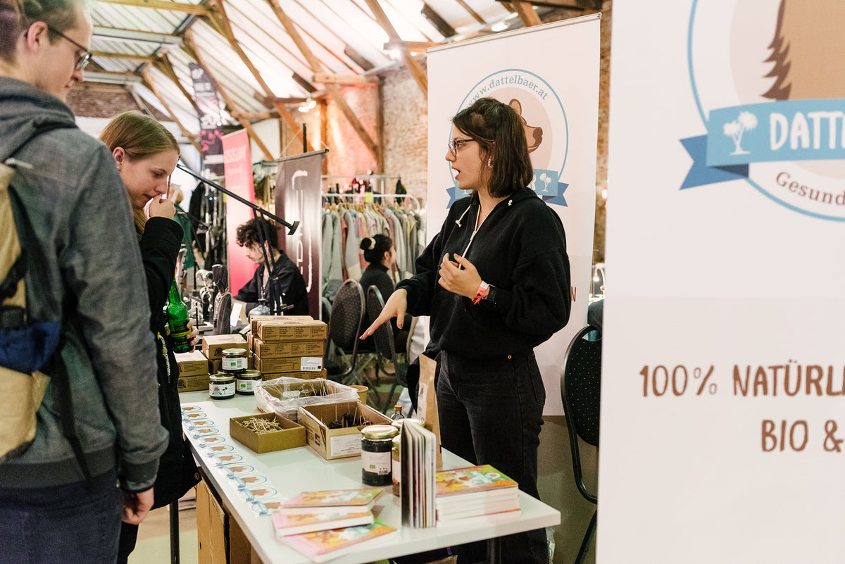 Eine Frau steht hinter einem Stand in einer Markthalle und spricht mit zwei Personen. Am Stand stehen Gläser, Proben und Broschüren. Schilder rund um den Stand werben für natürliche und biologische Produkte. Im Hintergrund sind andere Stände und Kunden zu sehen.