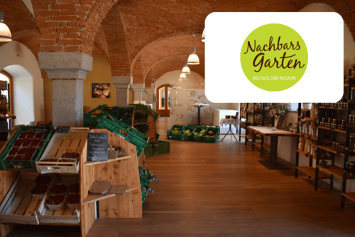 Ein rustikaler Biomarkt mit gewölbten Ziegeldecken, hölzernen Regalen mit Produkten und Waren, grünen Kisten und einem Logo mit der Aufschrift Nachbars Garten Bio aus der Region in der oberen rechten Ecke.
