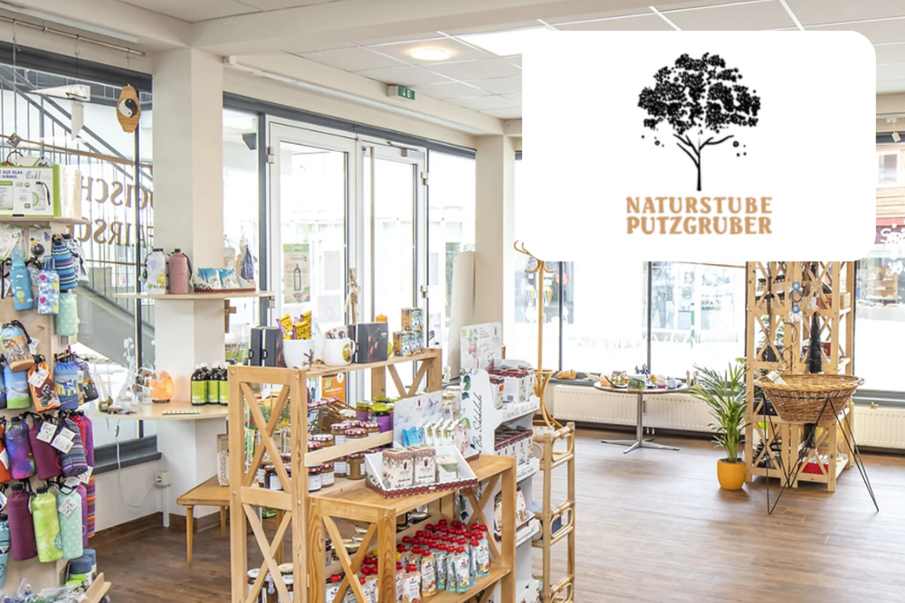 Ein helles, modernes Ladeninnere mit Holzregalen, in denen verschiedene Naturprodukte ausgestellt sind. Große Fenster lassen natürliches Licht herein. In der oberen rechten Ecke befindet sich ein Logo mit einem Baum und dem Text Naturstube Putzgruber.
