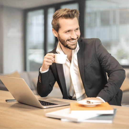 Ein lächelnder Mann im Anzug sitzt an einem Schreibtisch mit einem Laptop und hält eine Tasse Kaffee. Auf dem Schreibtisch liegen Papiere und ein Stift, und der Hintergrund des Büros wirkt hell und modern.