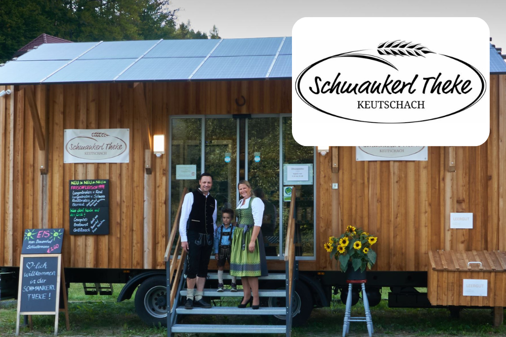 Eine Familie in traditioneller Kleidung steht am Eingang eines hölzernen Imbisswagens mit Sonnenblumen neben der Tür. Auf dem Schild steht Schwamkerl Theke Keutschach. Auf dem Dach befinden sich Sonnenkollektoren.