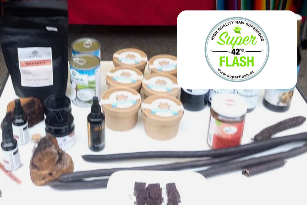 Auf einem Schautisch sind verschiedene Naturkostprodukte ausgestellt, darunter Gläser, Flaschen, Beutel und lange getrocknete Schoten. Eine weiße Karte zeigt das Super Flash-Logo mit dem Text High Quality Raw Superfood und die Website superflash.nl.