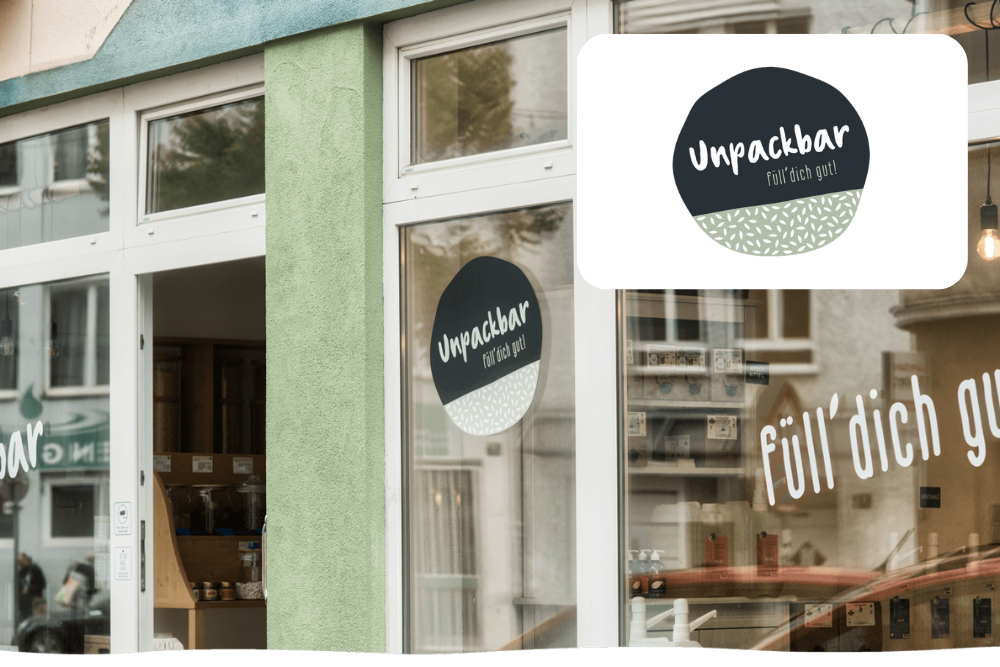 Schaufenster mit großen Fenstern, die das Logo Unpackbar Füll dich gut in einem kreisförmigen Design zeigen. Das Innere zeigt Holzregale mit Produkten, die auf einen Zero-Waste- oder Nachfüll-Laden hindeuten.