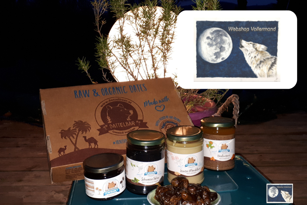 Eine Auslage mit Naturprodukten auf einem Tisch umfasst Gläser mit Brotaufstrichen, eine Schachtel mit der Aufschrift RAW & ORGANIC DATES und eine Schale mit Datteln. Im Hintergrund sind Pflanzen und eine helle Lampe zu sehen. Ein Einsatz zeigt einen Wolf, der den Vollmond anheult.