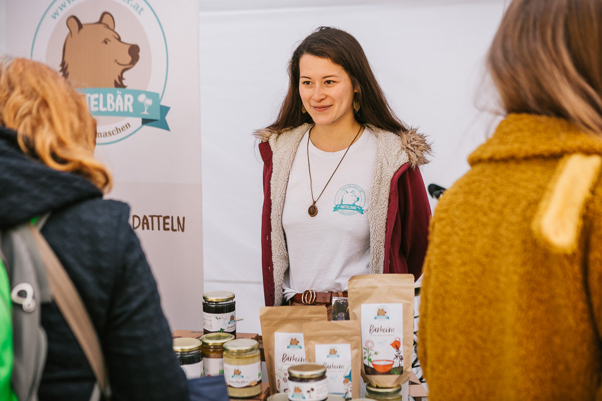 Eine Frau steht hinter einem Tisch, der Gläser und Tüten mit Lebensmitteln an einem Marktstand ausstellt. Sie lächelt zwei Personen an, die vor dem Stand stehen, der ein Bärenlogo auf einem Transparent trägt.