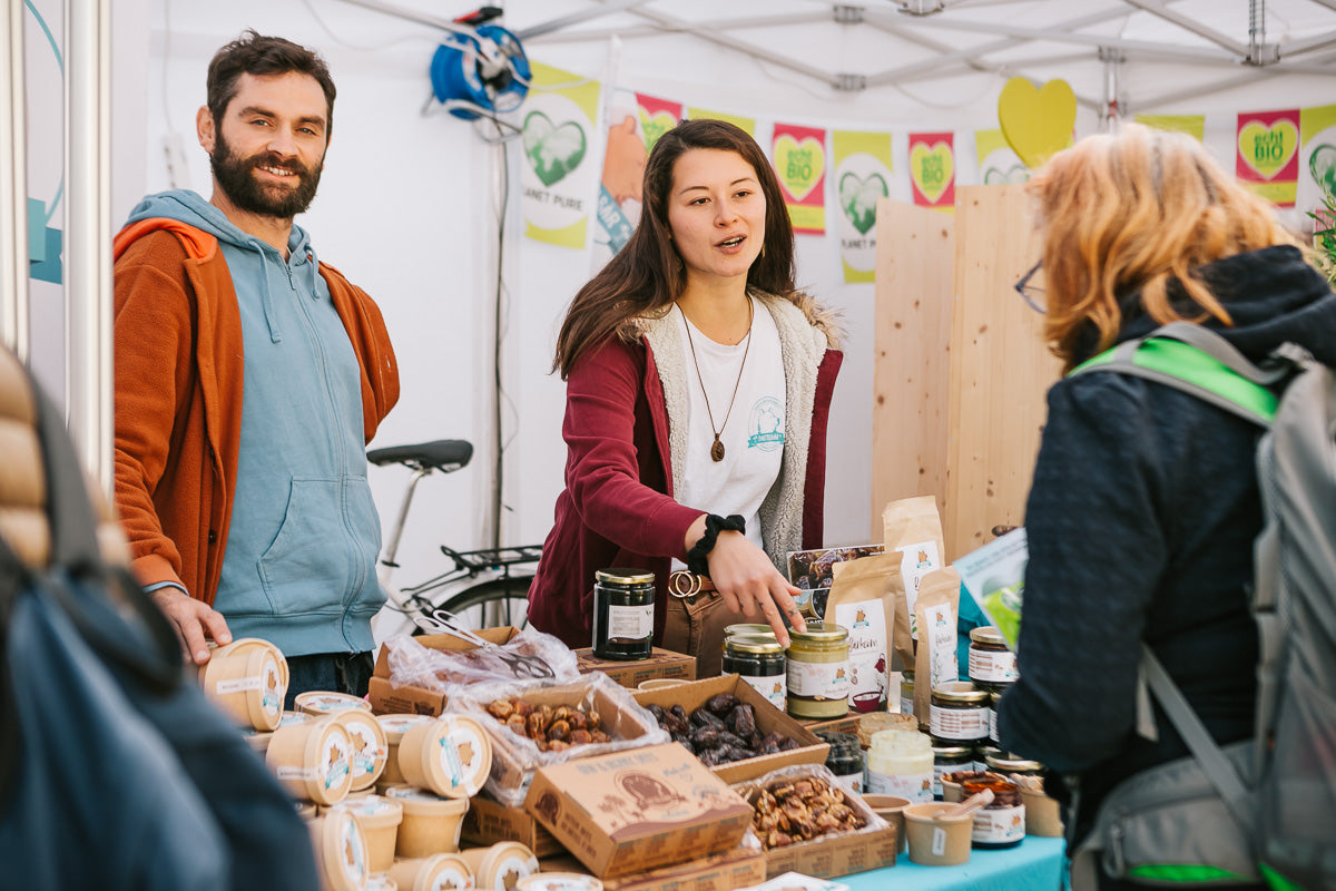 Zwei Verkäufer stehen hinter einem Marktstand mit verschiedenen verpackten Lebensmitteln, Gläsern und Snacks und unterhalten sich mit einem Kunden. Im Hintergrund sind bunte Werbebanner und Produktauslagen zu sehen.