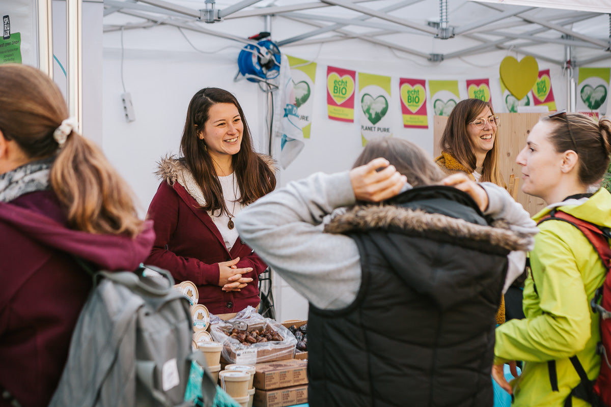 Eine Frau lächelt, während sie mit Kunden an einem Marktstand spricht, der Bioprodukte verkauft. Andere Frauen stehen in der Nähe, und im Hintergrund sind Schilder mit grünen Herzlogos und dem Wort "BIO" zu sehen.