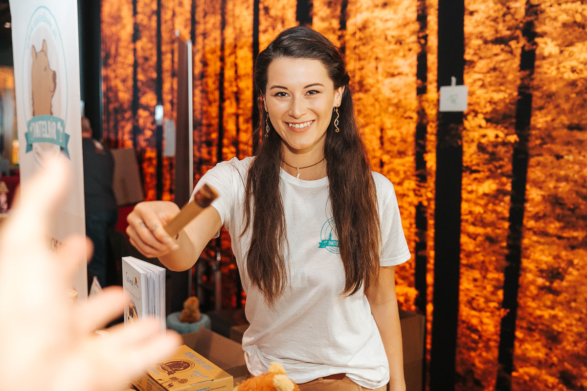 Eine lächelnde Frau mit langen braunen Haaren, die ein weißes T-Shirt trägt, streckt jemandem im Vordergrund einen kleinen Gegenstand entgegen. Sie steht an einem Stand mit warmen orangefarbenen Lichtern und einem Hintergrund mit Naturmotiven.