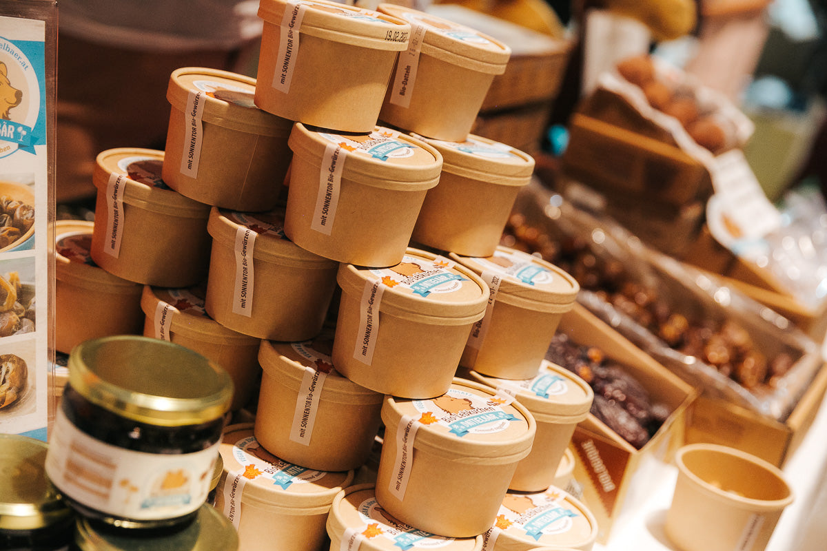 Ein pyramidenförmiger Stapel brauner Papierbecher mit blauen und weißen Etiketten an einem Marktstand, umgeben von Gläsern und anderen verpackten Lebensmitteln. Die Becher scheinen zum Verkauf angebotene Lebensmittel zu enthalten.