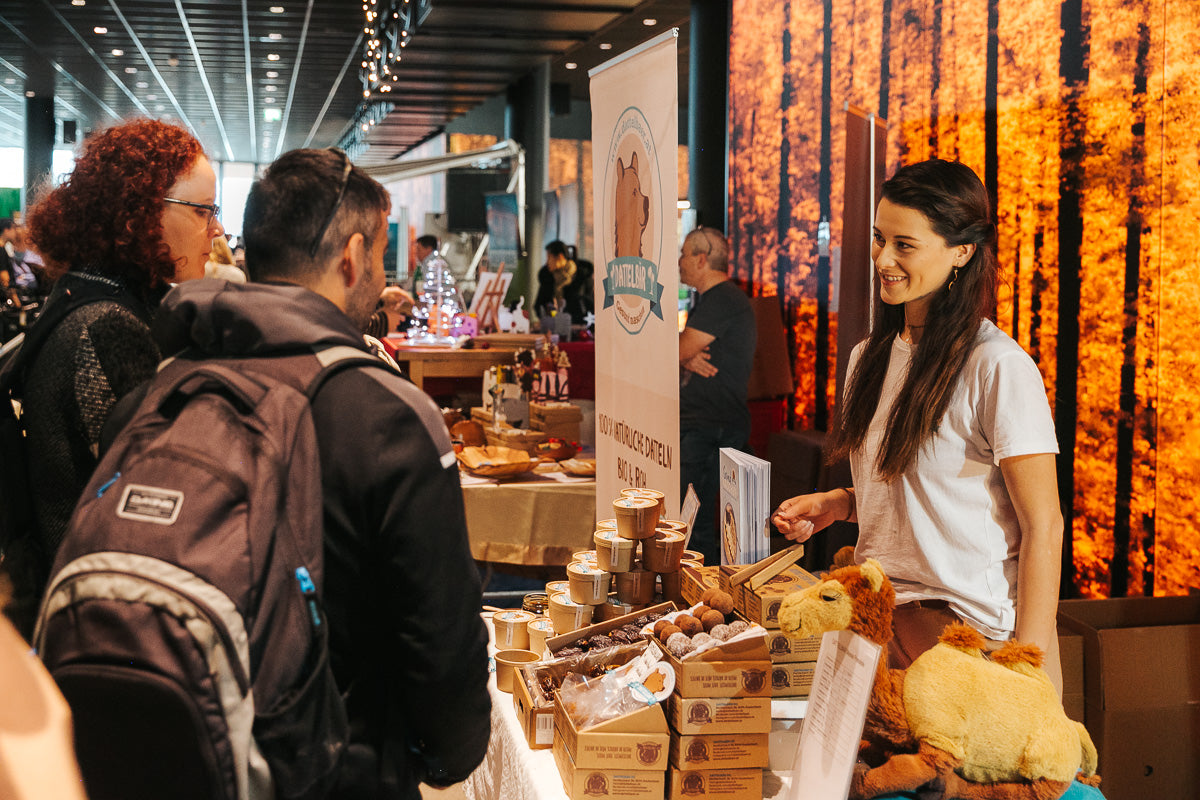 Eine Frau steht hinter einem Marktstand mit Produkten rund um das Kamel, lächelt und spricht mit zwei Kunden. Die Auslage umfasst Dosen, Gläser und ein Kamel-Plüschtier. Im Hintergrund sind andere Stände und warme Beleuchtung zu sehen.