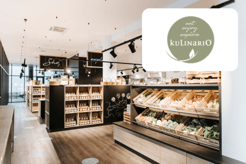 Modernes Lebensmittelgeschäft mit Holzregalen, in denen frische Produkte und verpackte Waren ausgestellt sind; in der Ecke befindet sich ein großes Logo mit der Aufschrift eat enjoy explore KuLinariO und einem grünen Blattmuster.